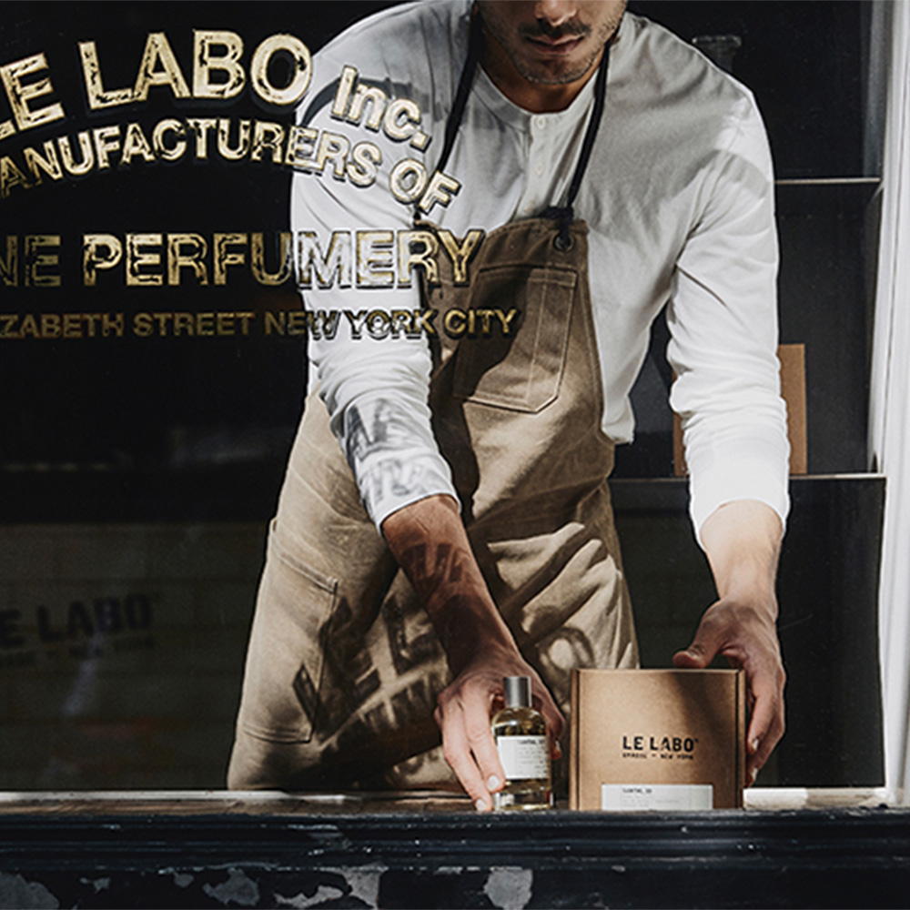 Man in white shirt and brown apron arranging fragrance bottles and packaging on display shelf with gold branding text