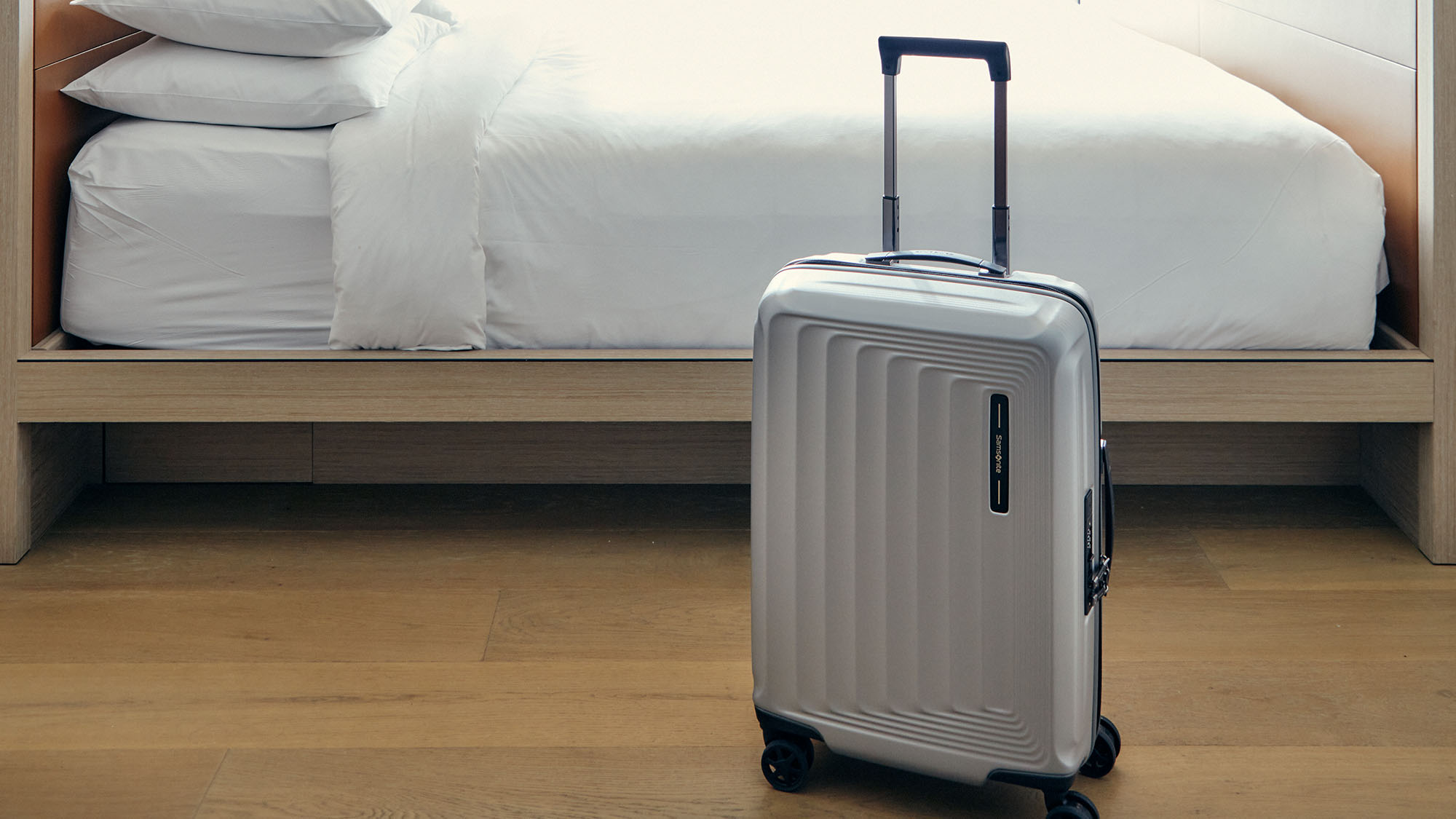 Silver hard-shell cabin suitcase with retractable handle and wheels positioned in front of a modern bed with white pillows and bedding