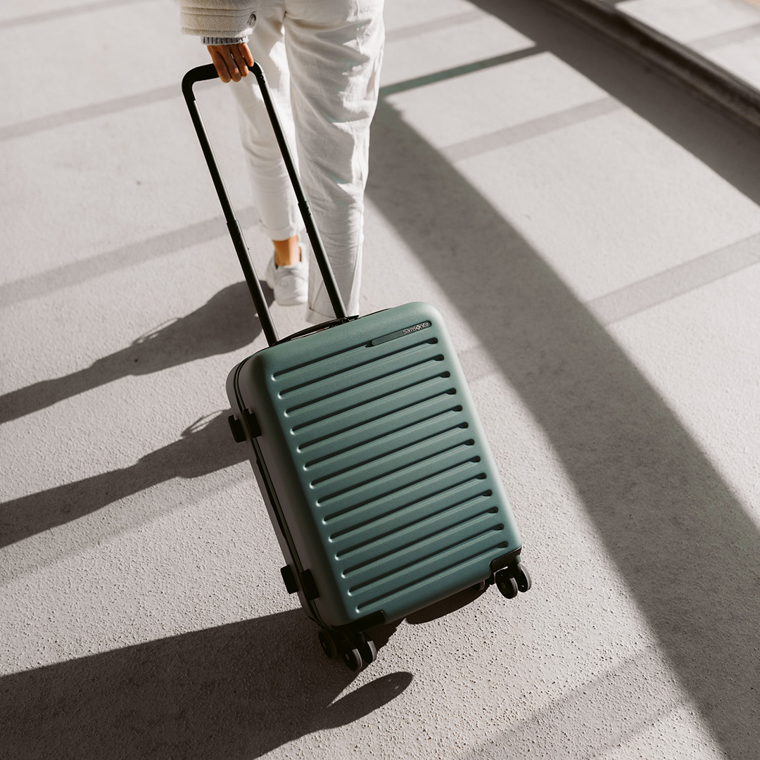 Person in white trousers and beige jumper pulling a sage green lightweight cabin suitcase across a bright tiled floor