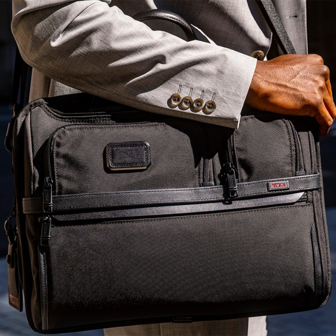 Man wearing tailored grey blazer and light blue shirt holding a charcoal grey business briefcase with leather trim