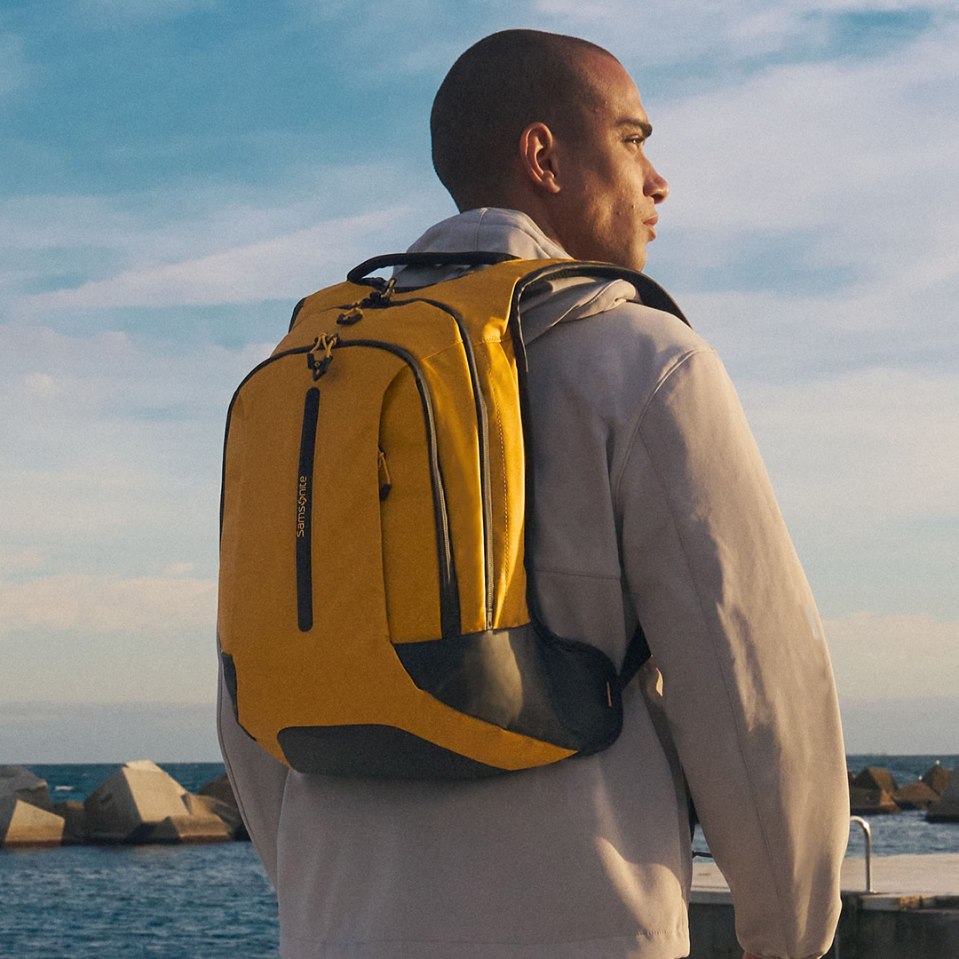 Man wearing grey jacket standing by coastal waterfront wearing a mustard yellow and navy backpack over his shoulder