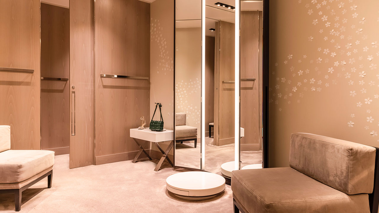 A luxurious fitting room with seating, mirrors, floral wall patterns, and a small table with a handbag.