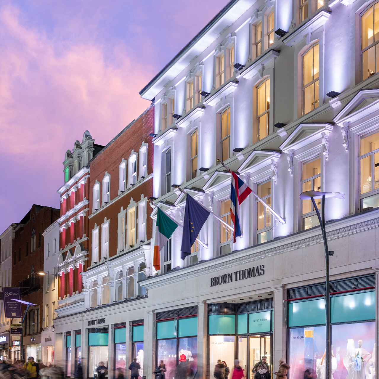 Illuminated storefront of Brown Thomas in Dublin, with flags and a pink sky at dusk..

