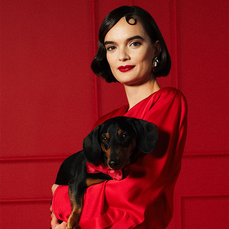 Woman in a red dress holding a small black and brown dog against a red background.