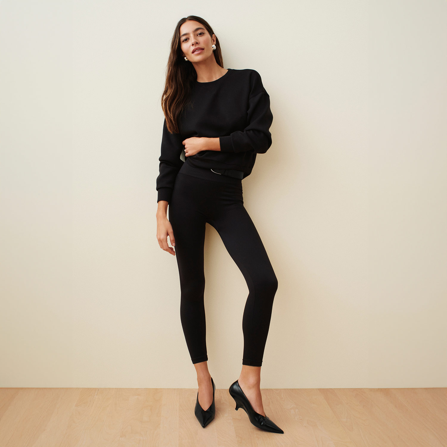 A woman in all-black attire poses against a neutral wall, wearing a sweater, leggings, and pointed shoes.
