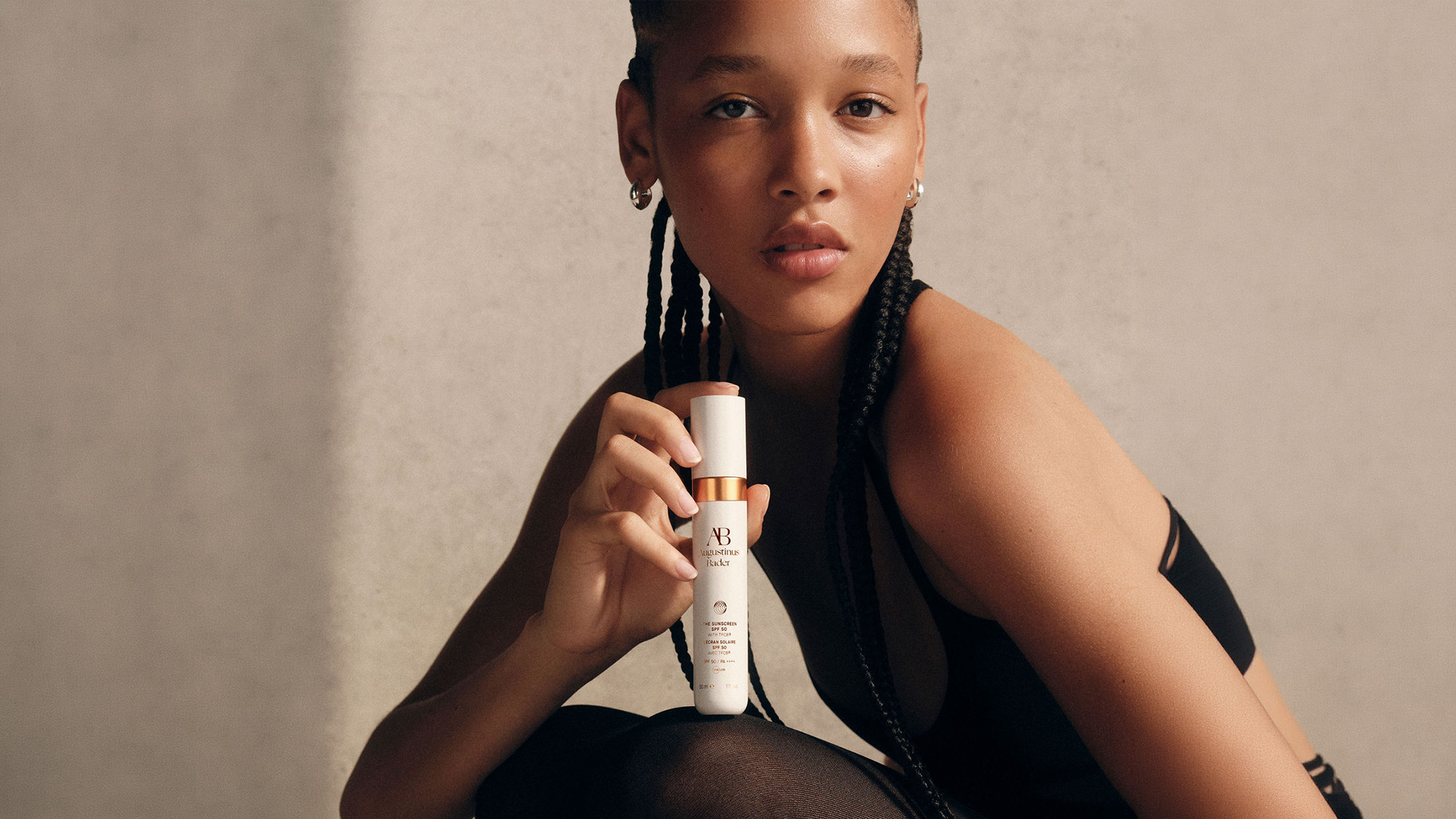 Woman holding white SPF sunscreen bottle with gold accents, wearing black sleeveless top against neutral background