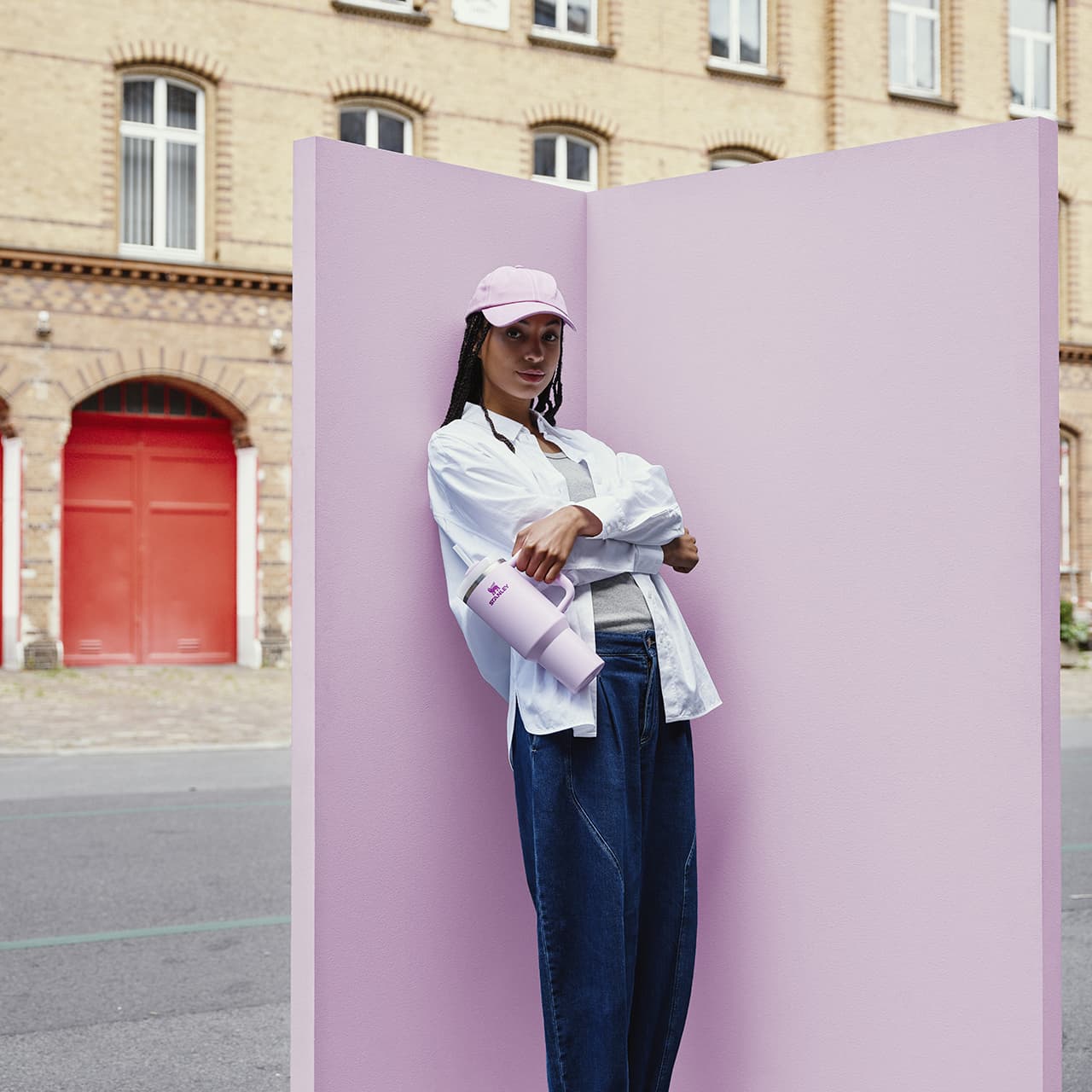 Woman wearing pink cap and white shirt holding lavender Stanley cup against pink wall
