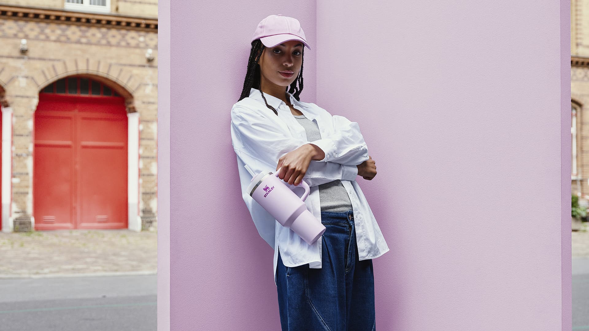 Woman wearing pink cap and white shirt holding lilac Stanley water bottle against pink wall