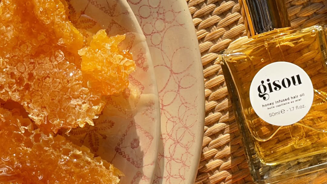 Honey-infused hair oil in glass bottle next to golden hair treatment texture in white bowl on woven surface