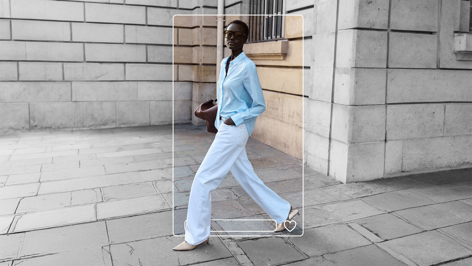 Woman wearing light blue shirt and white trousers with brown handbag, standing against stone building.