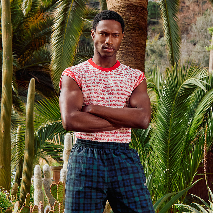A person stands with crossed arms in a lush garden filled with cacti and palm trees.