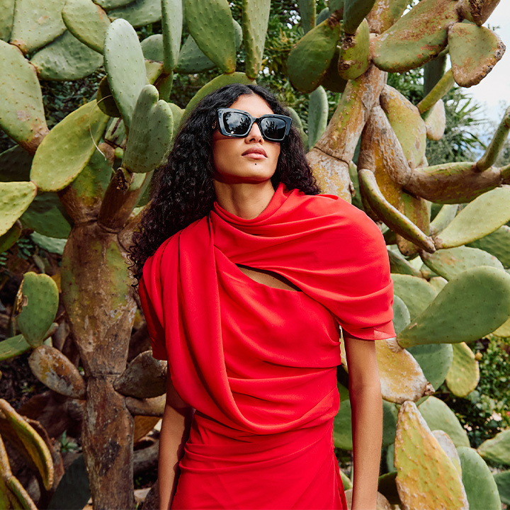 A person in a vibrant red dress and sunglasses stands in front of large, green cactus plants.