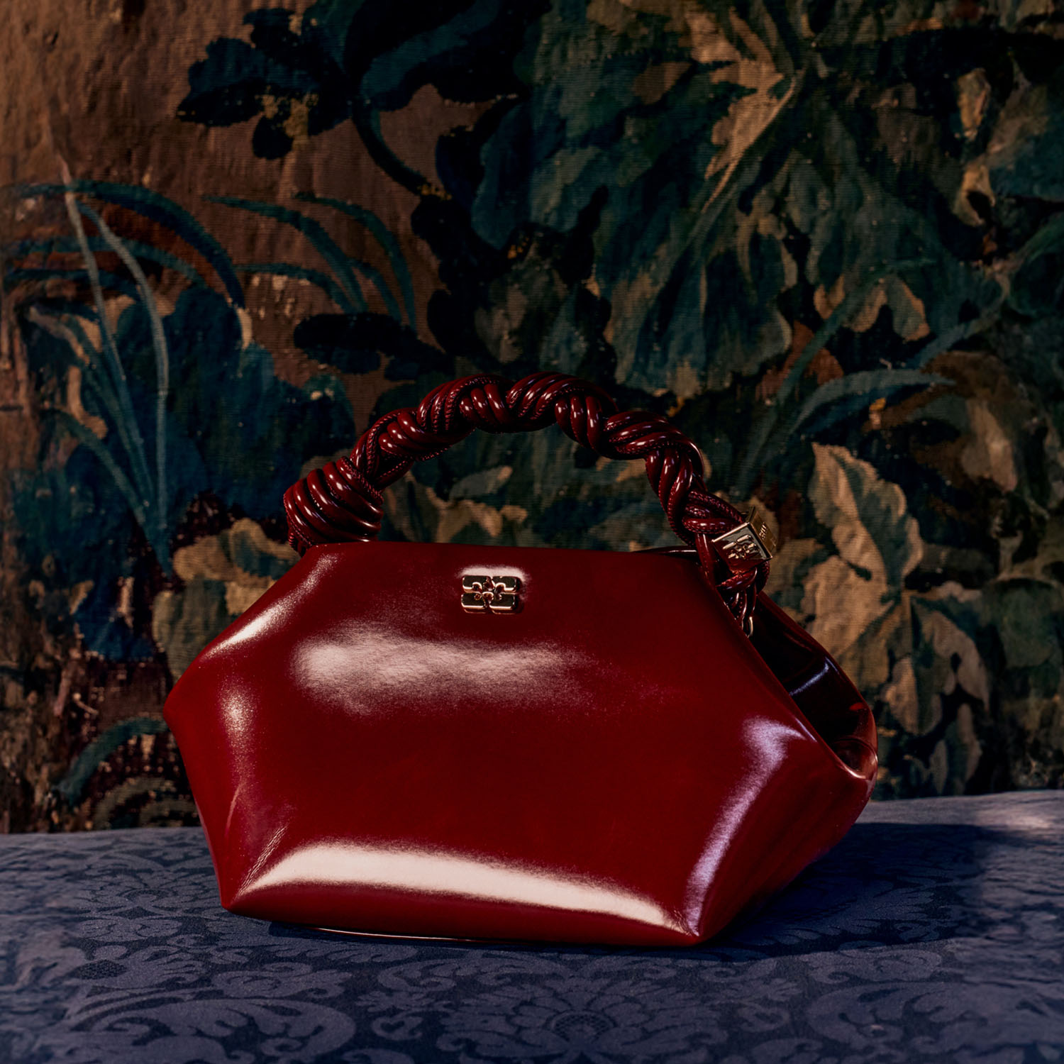 A shiny red handbag with a twisted handle sits against a dark floral tapestry background.