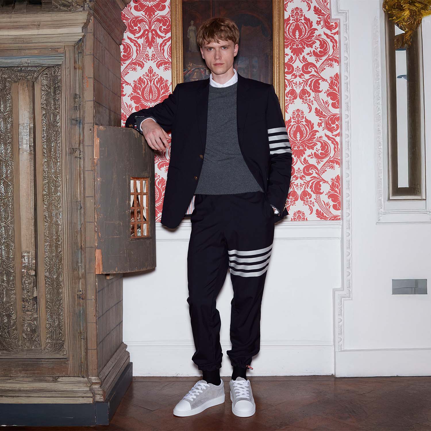 A person stands in front of a detailed tapestry, wearing a fuzzy grey sweater and dark pants.