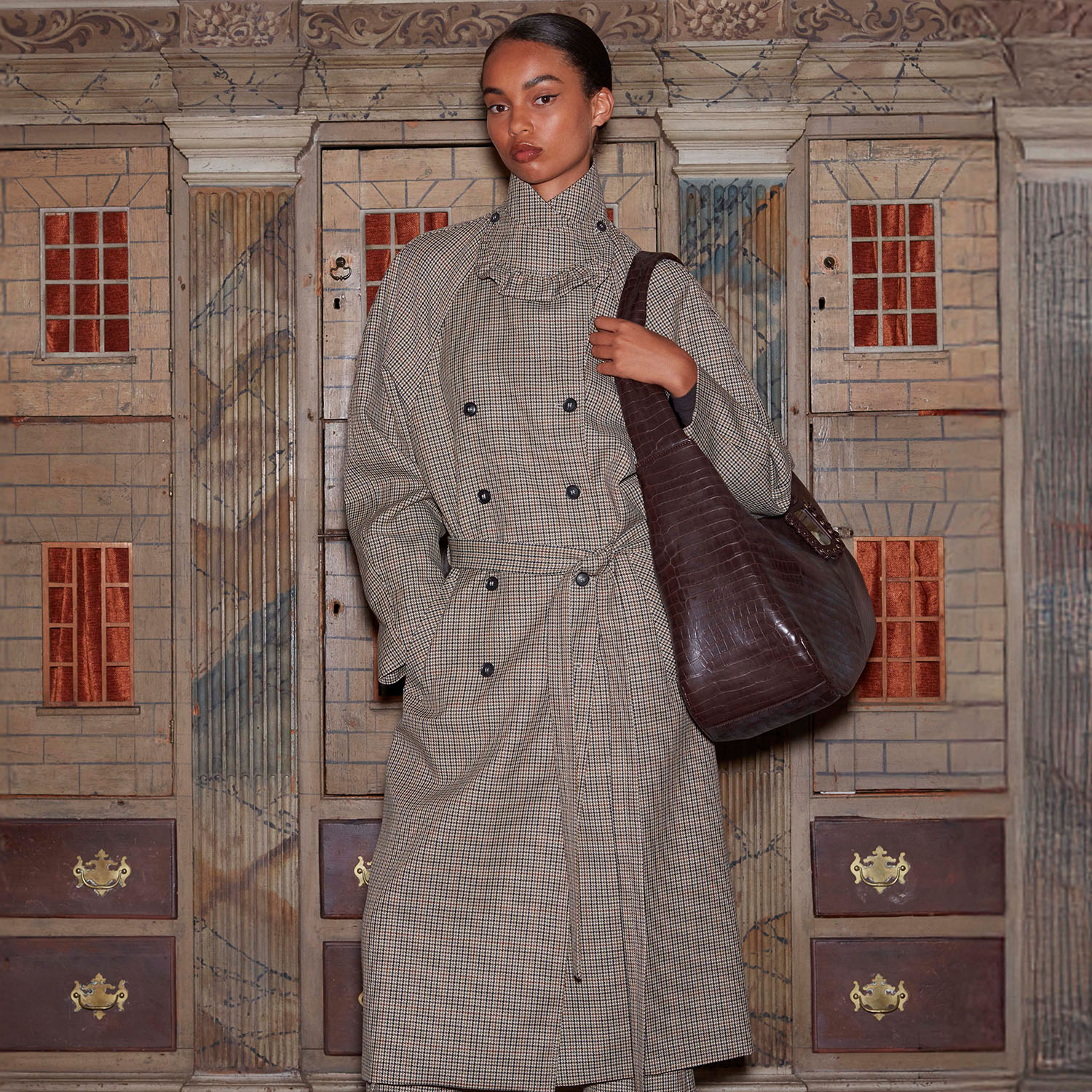 A person in a plaid trench coat and pants stands with a large brown bag, against an ornate, architectural backdrop with red accents.