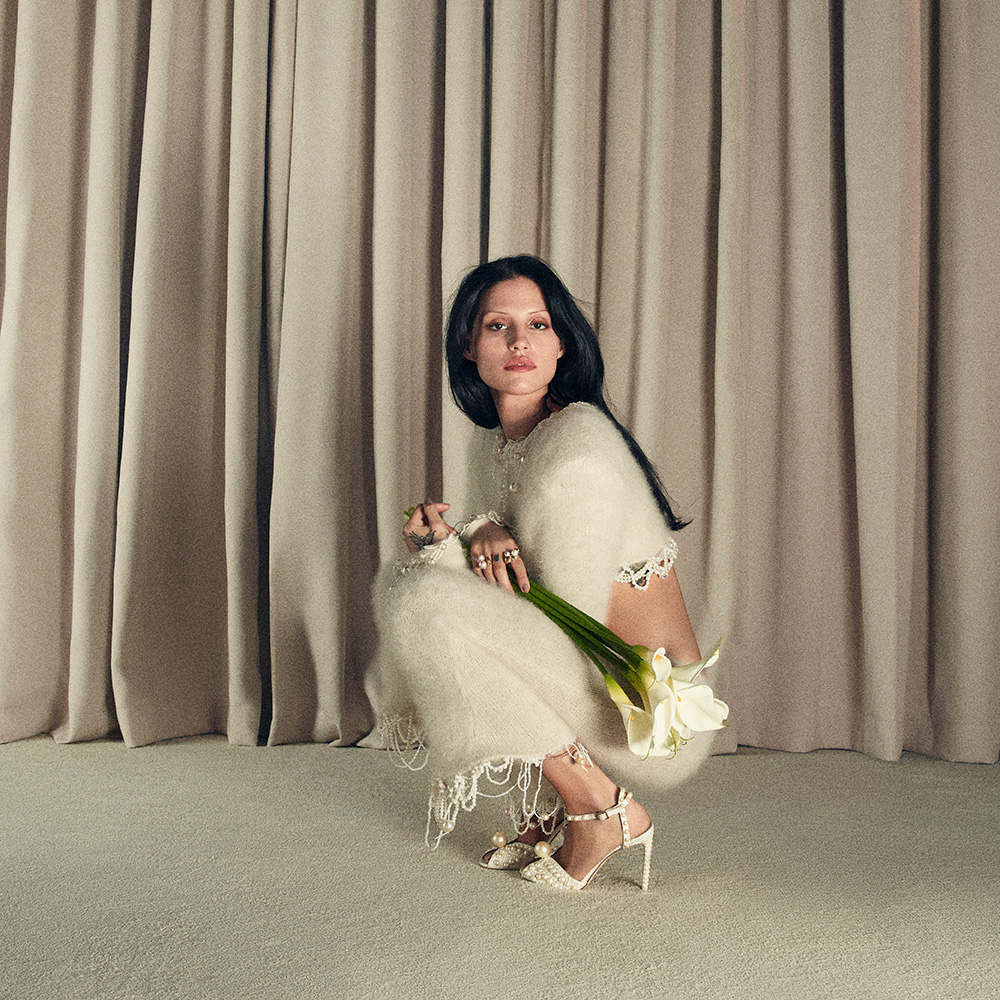 A person squats on a carpeted floor wearing a white outfit, holding flowers, with a curtain backdrop.