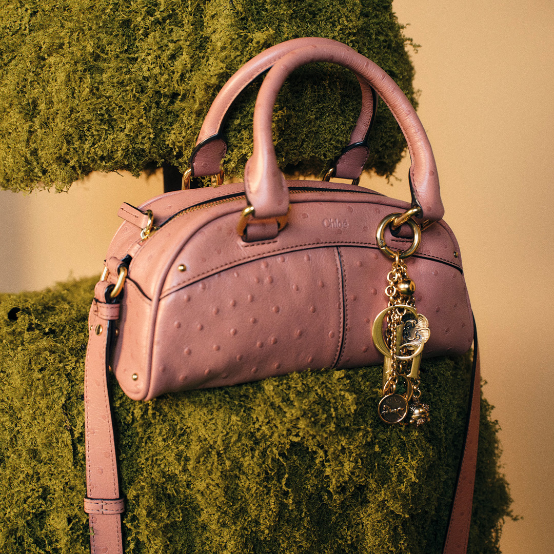A pink handbag with gold charms hangs on a green moss-covered surface.