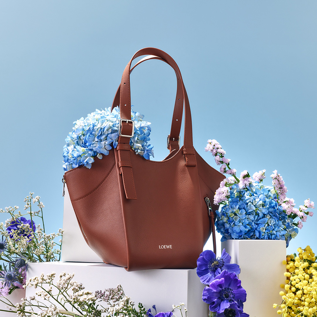 A brown handbag surrounded by an arrangement of colorful flowers against a light blue background.