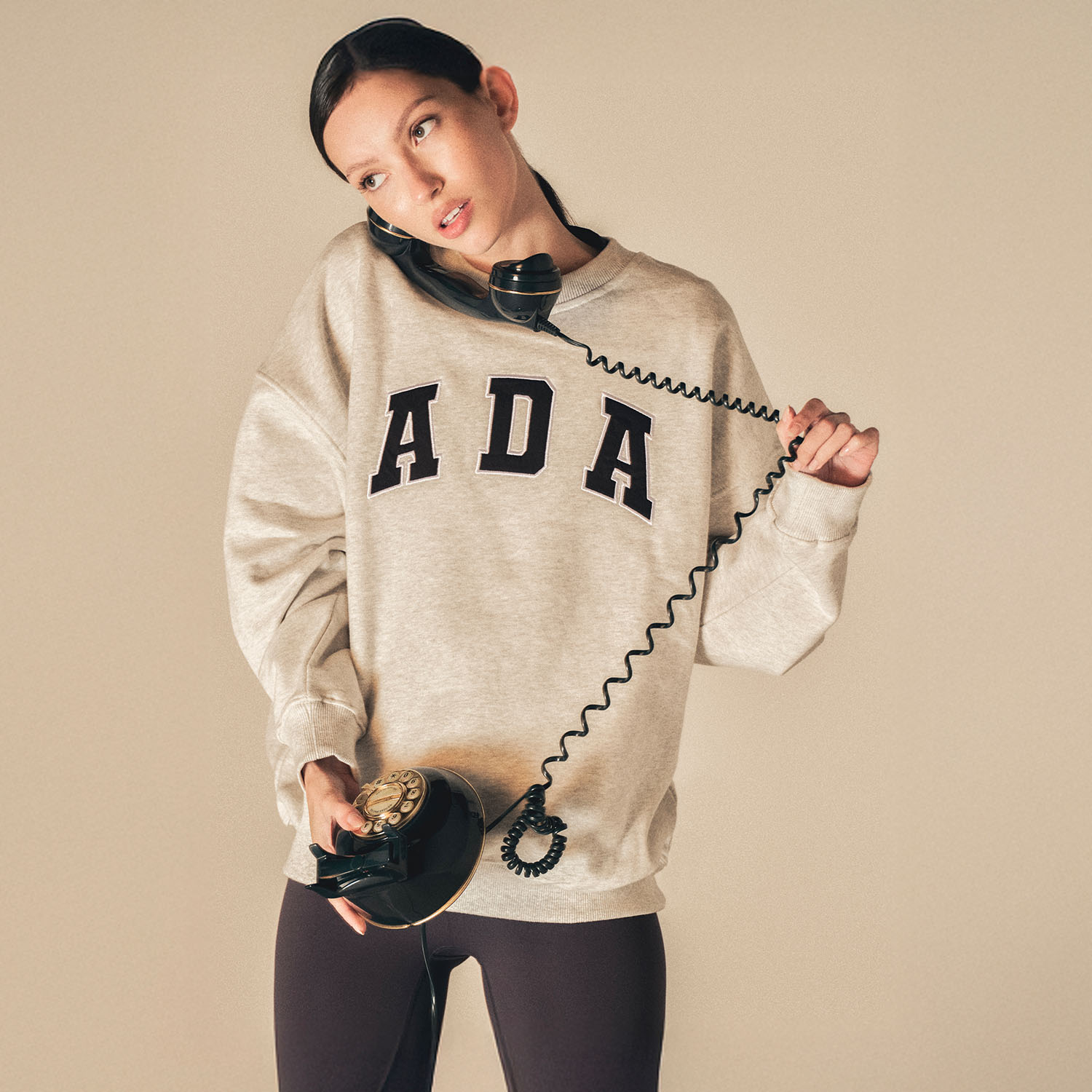Person in a gray 'ADA' sweatshirt uses a vintage rotary phone, tilting their head to hold the receiver.