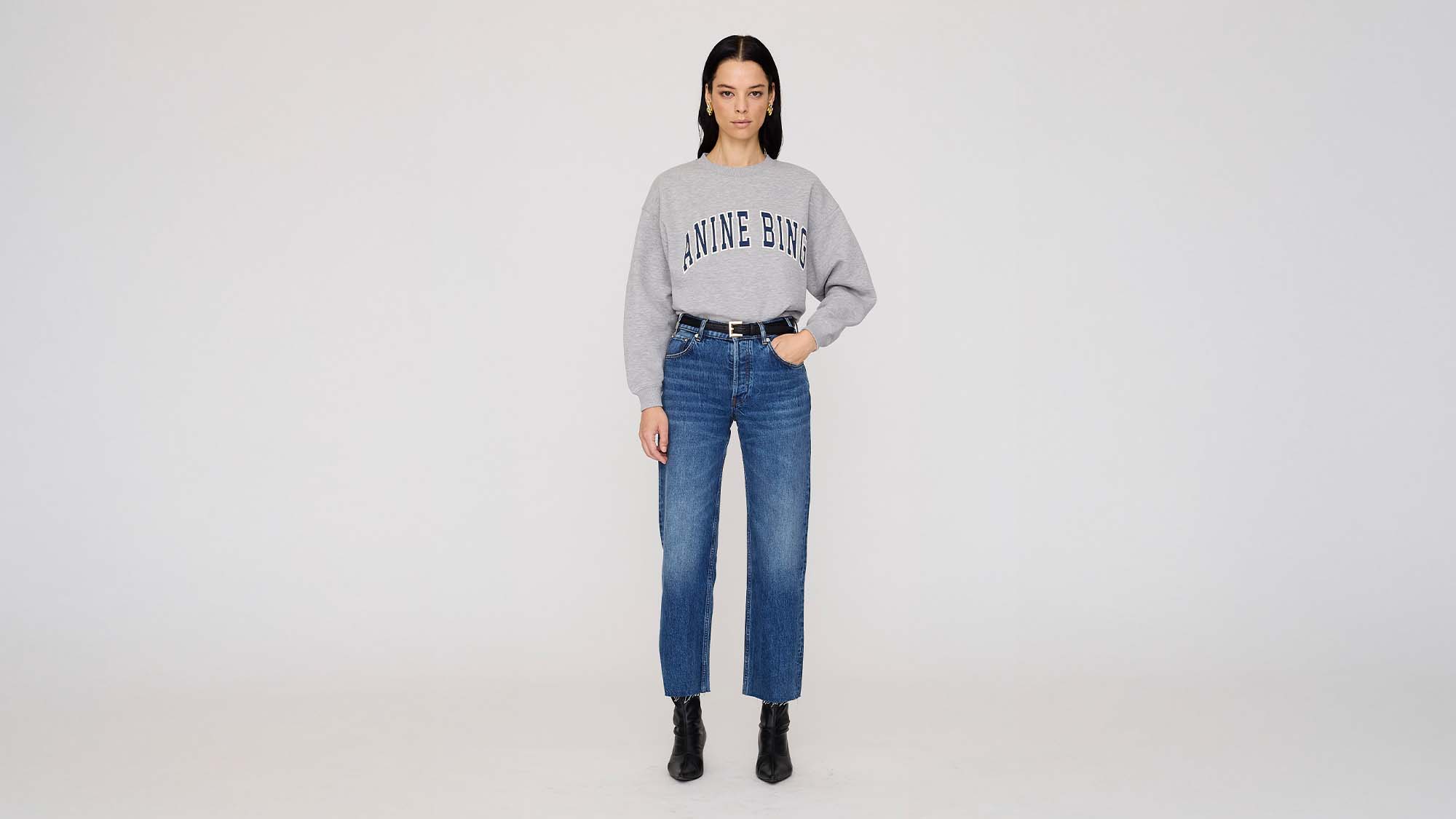 A woman stands wearing a gray sweatshirt with 'ANINE BING' text, blue jeans, and black boots.