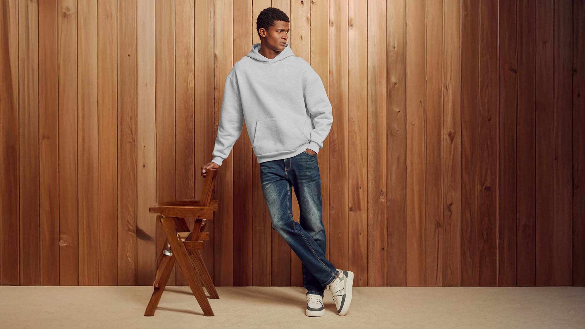 A person in a light gray hoodie and jeans leans against a wooden chair in front of a wooden panel wall.