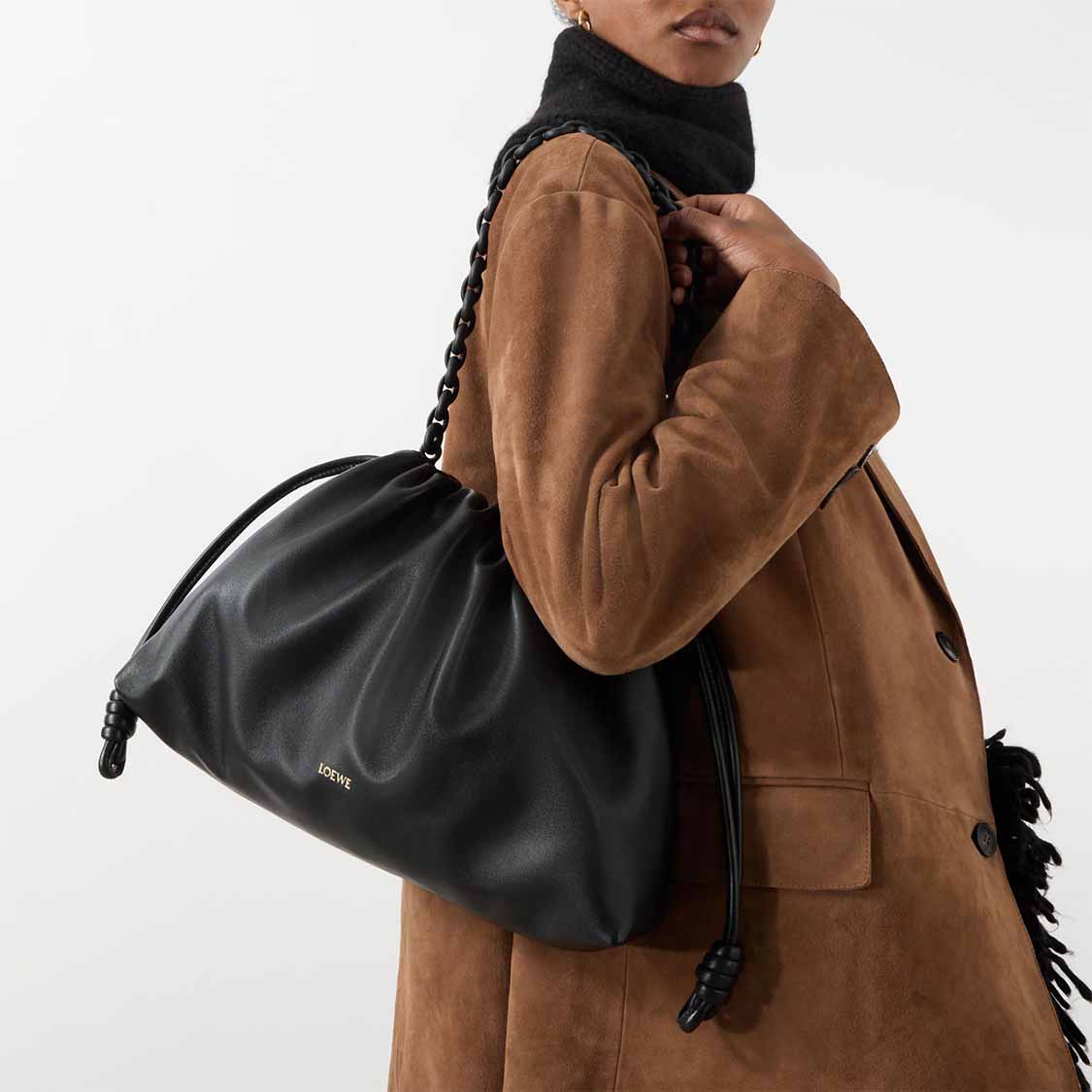 Person in a brown coat holding a large black Loewe bag with a knotted strap.
