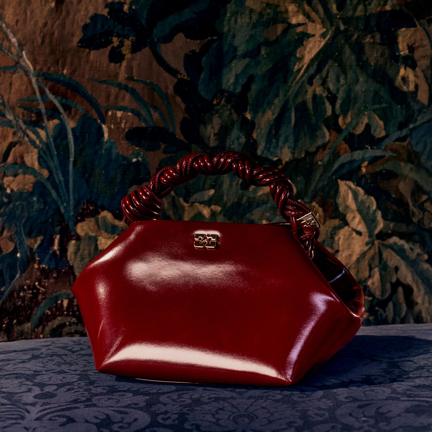 A shiny red handbag with a braided handle sits on a patterned surface against a floral tapestry background.