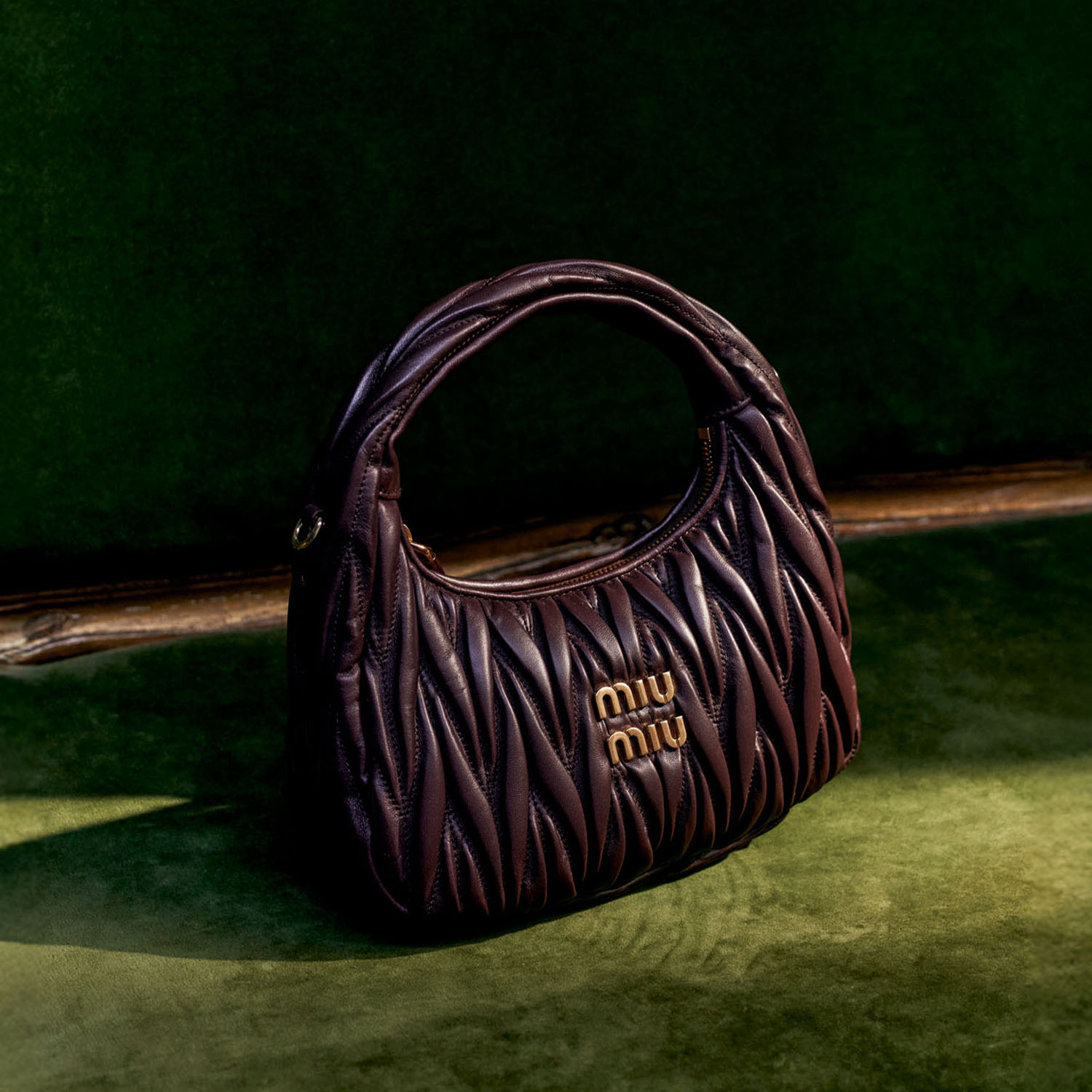 A dark brown quilted handbag with a rounded handle sits on a green surface, featuring a gold 'miumiu' logo.