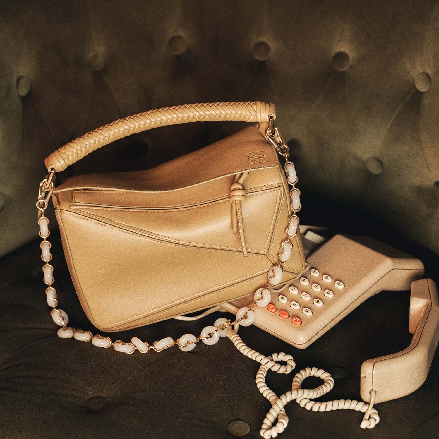 A beige handbag with a braided handle and bead strap sits on a green tufted chair beside a vintage beige corded phone.