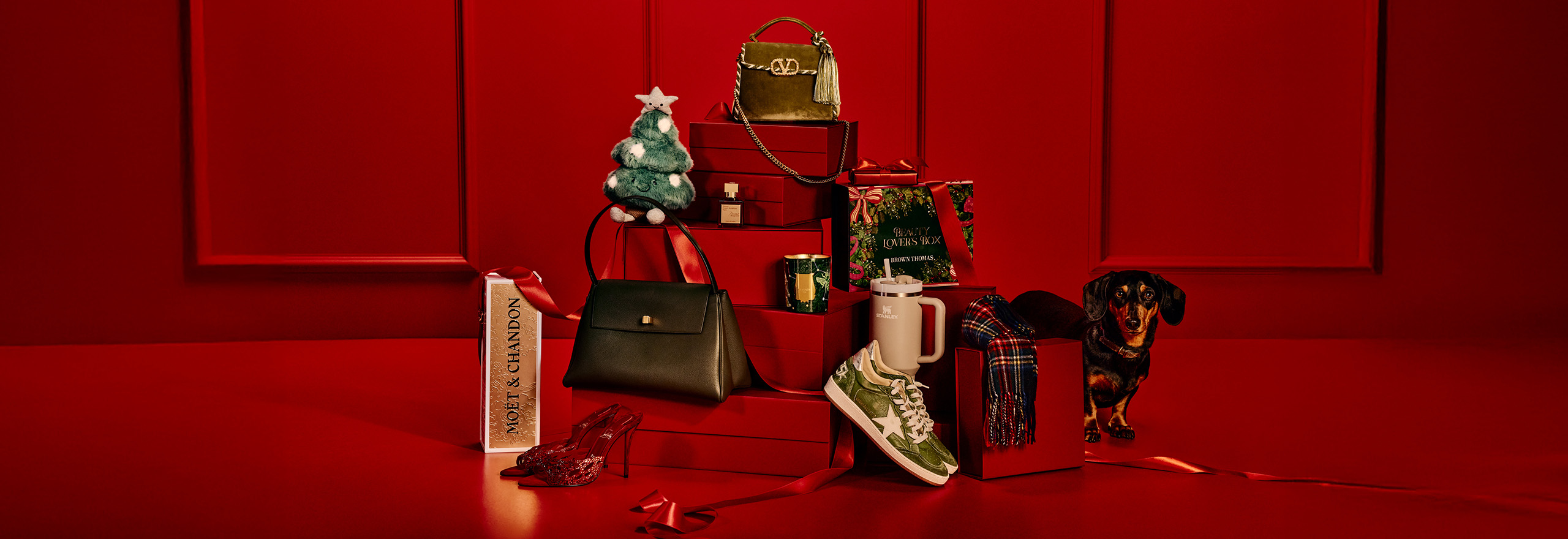 A festive display features luxury handbags, sneakers, a small Christmas tree, a gift box, and a dachshund on a red background.