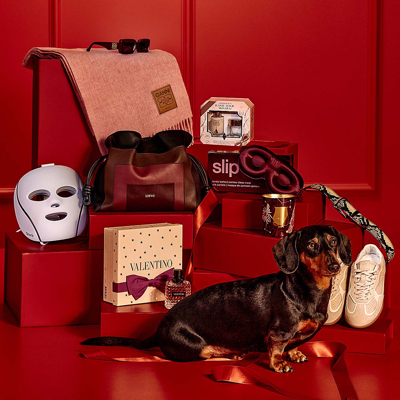 A dachshund stands among luxury items, including a face mask, perfume, sneakers, bag, and stacked red gift boxes against a red backdrop.