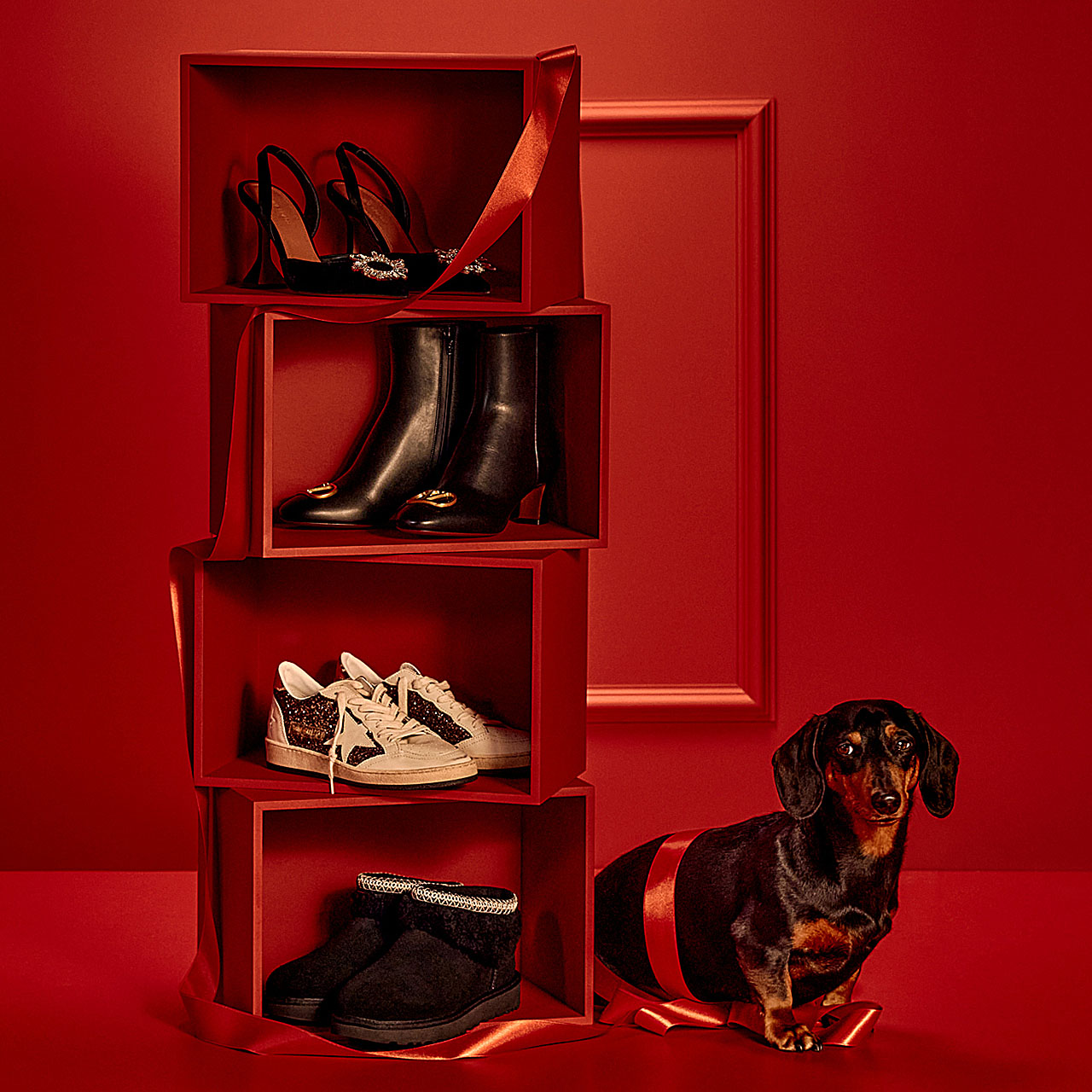 Red display with various shoes on shelves and a small black dog with a red ribbon standing beside it.