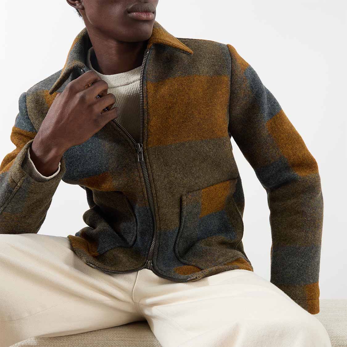 Person wearing a brown and gray plaid jacket over a white outfit, seated with hand near their face.