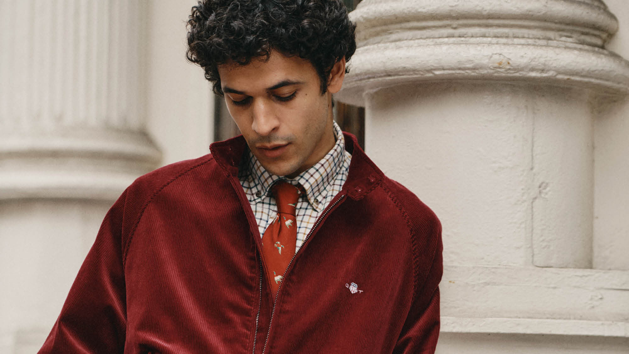 A man in a red jacket over a plaid shirt and tie stands by a white column, looking down.