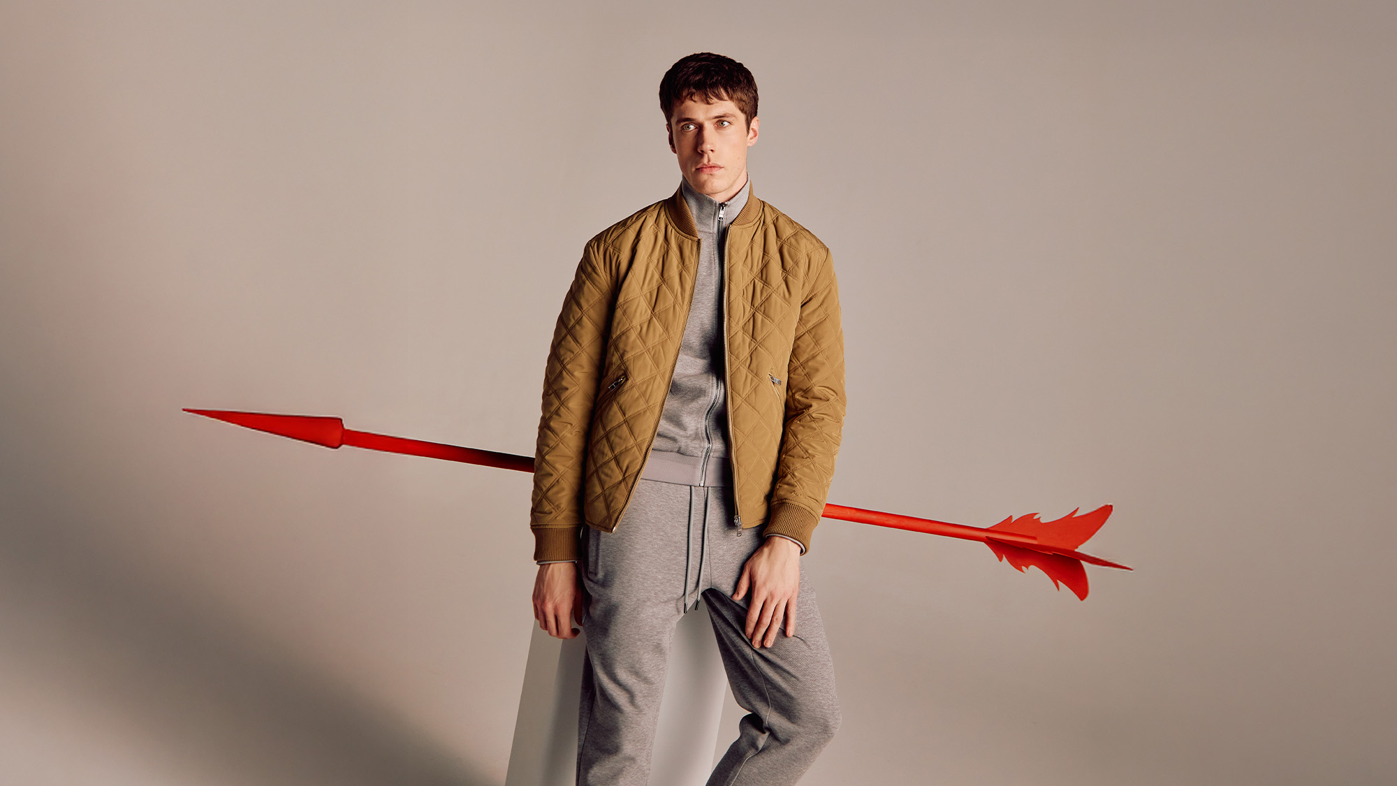 Man in a brown quilted jacket and gray tracksuit leans against a white block, with a large red arrow in the background.