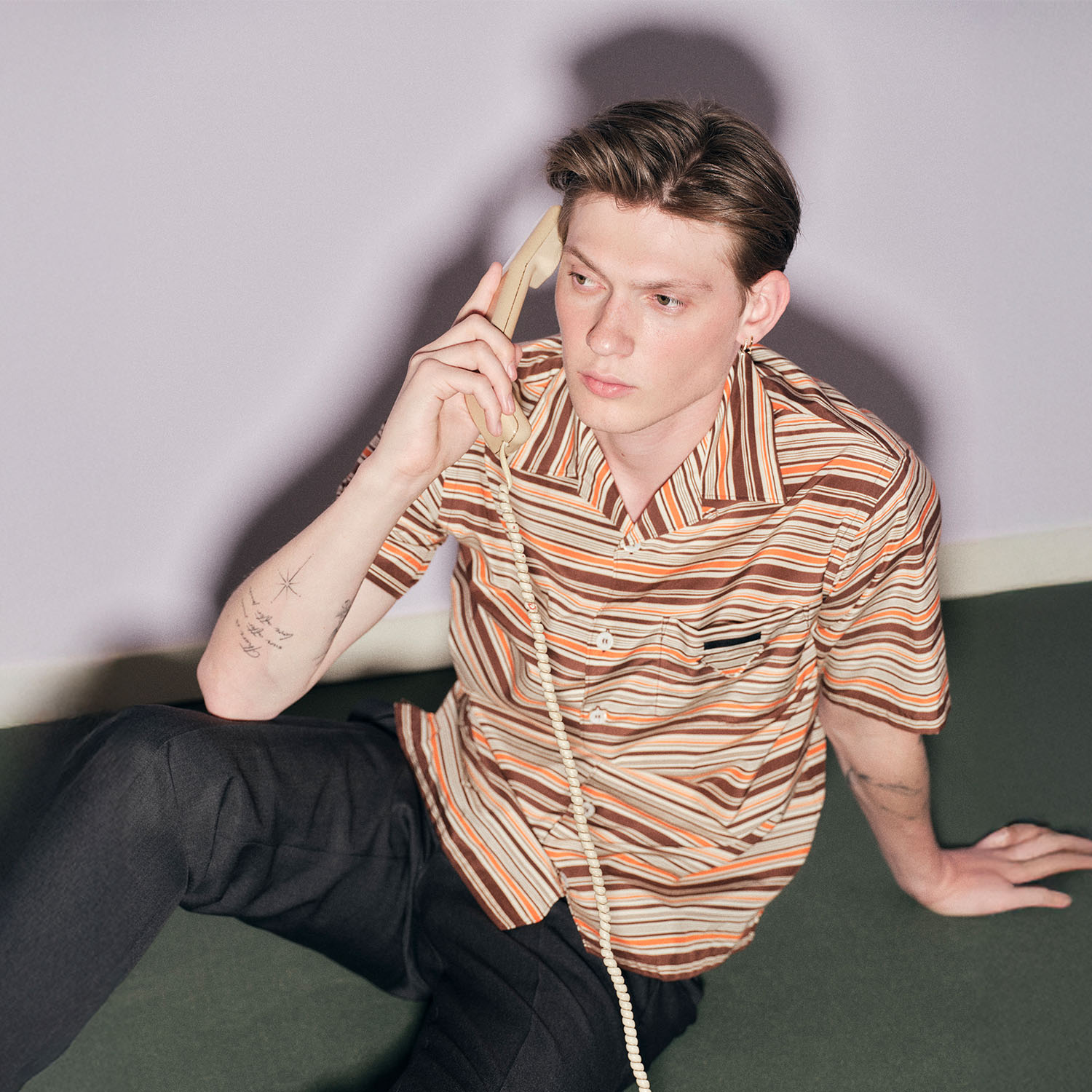 A person with tattoos sits on the floor, holding a rotary phone. They're wearing a striped shirt and dark pants, looking slightly away.