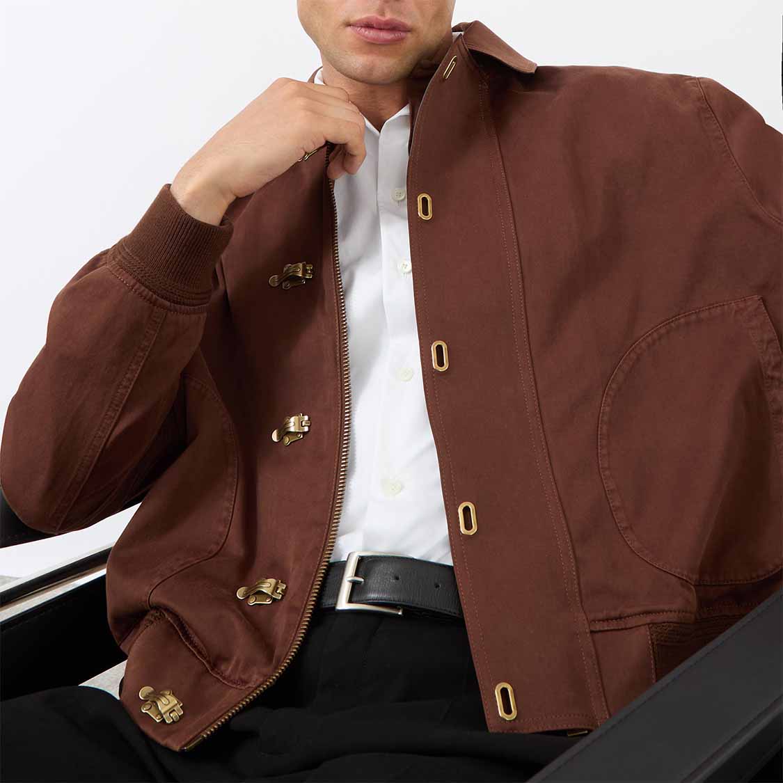Person seated, wearing a brown jacket with button details over a white shirt.