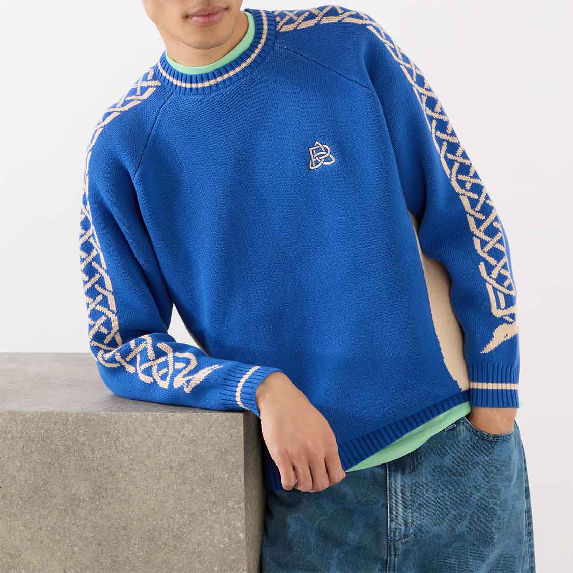A person wearing a blue sweater with patterned sleeves leans against a concrete block.