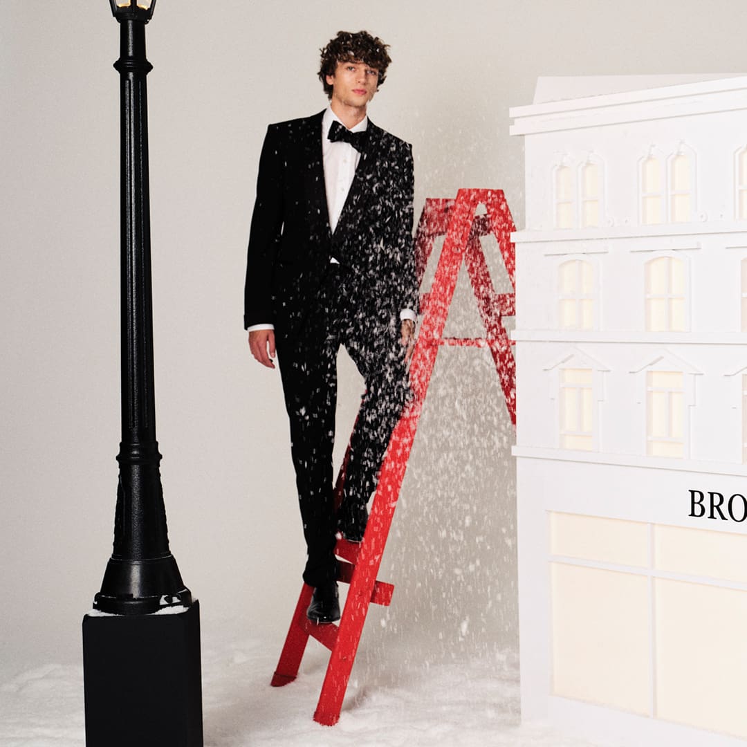 A person in a tuxedo stands on a red ladder surrounded by artificial snow, near a streetlamp and a white building facade.