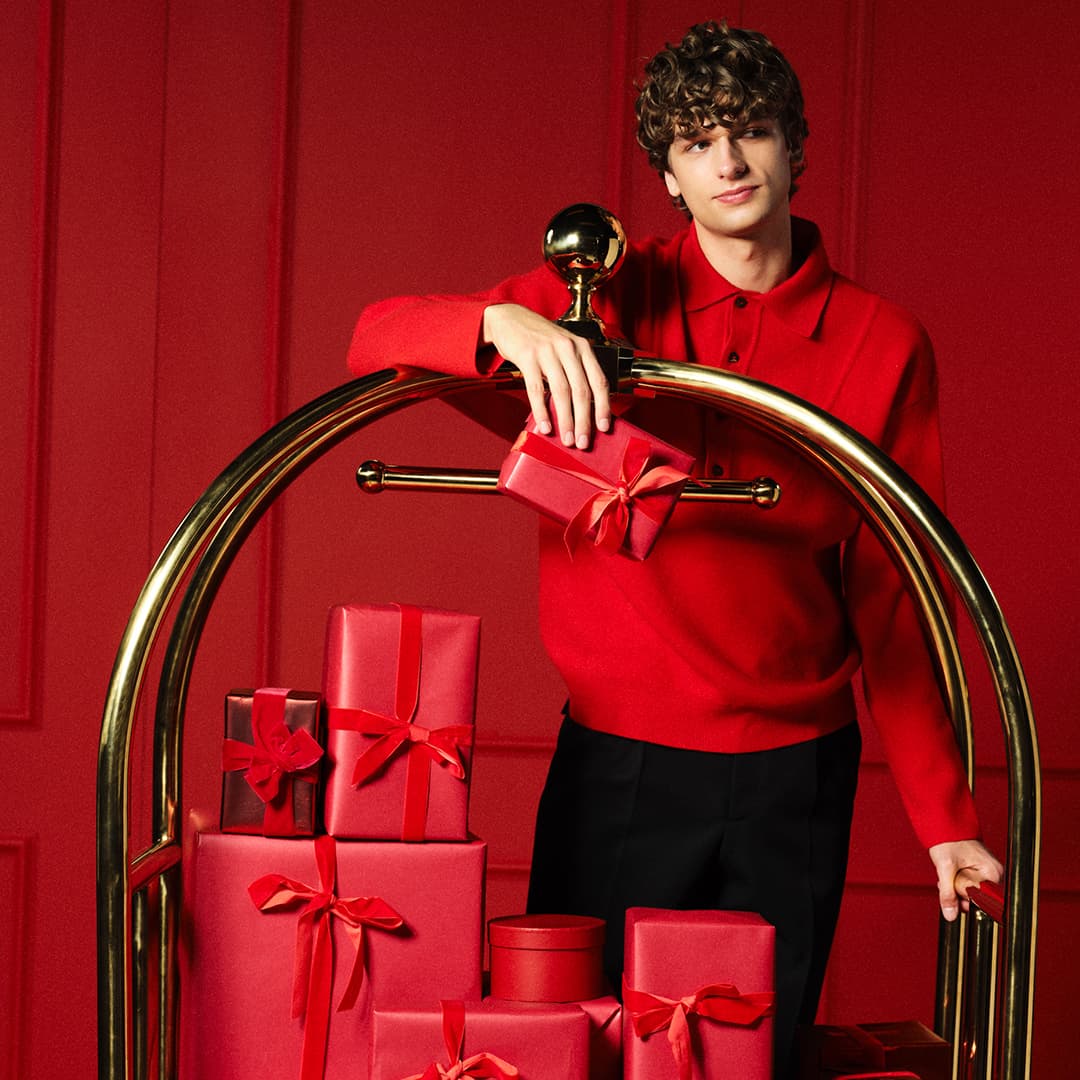A man in a red jumper leaning over a luggage trolly stacked with red gift boxes
