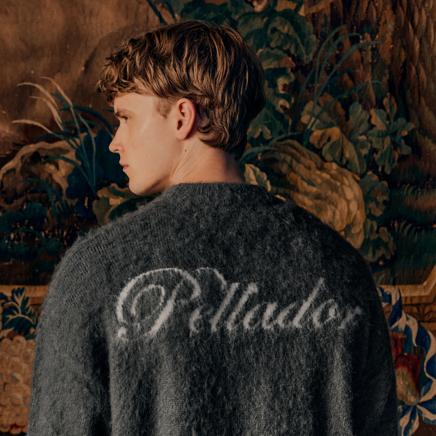 A person in a sweater with 'Peludo' on the back stands before a tapestry of a lion and nature scene.