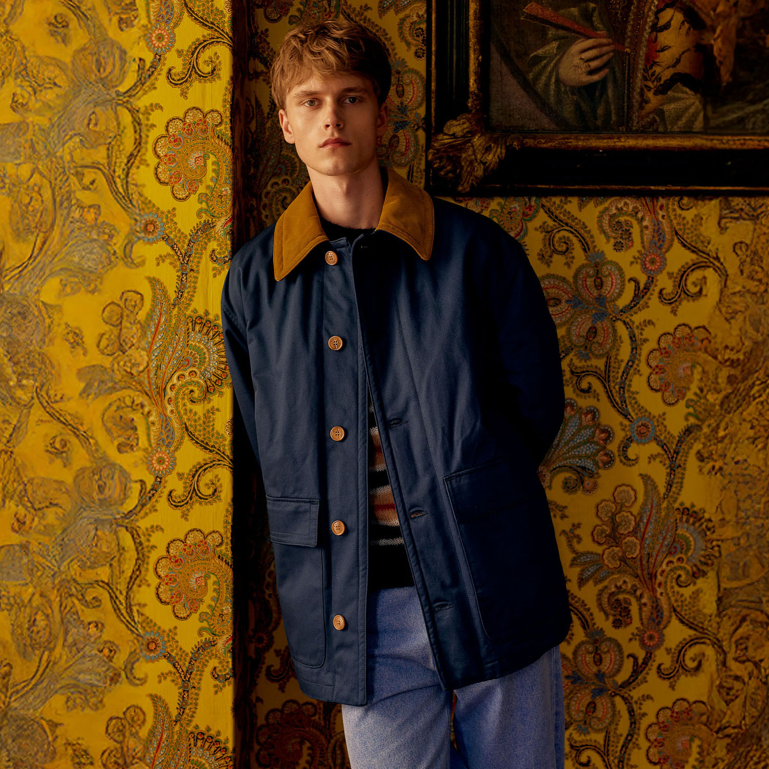 Man in a navy coat with a tan collar stands against a yellow, patterned wallpaper, near a framed painting.