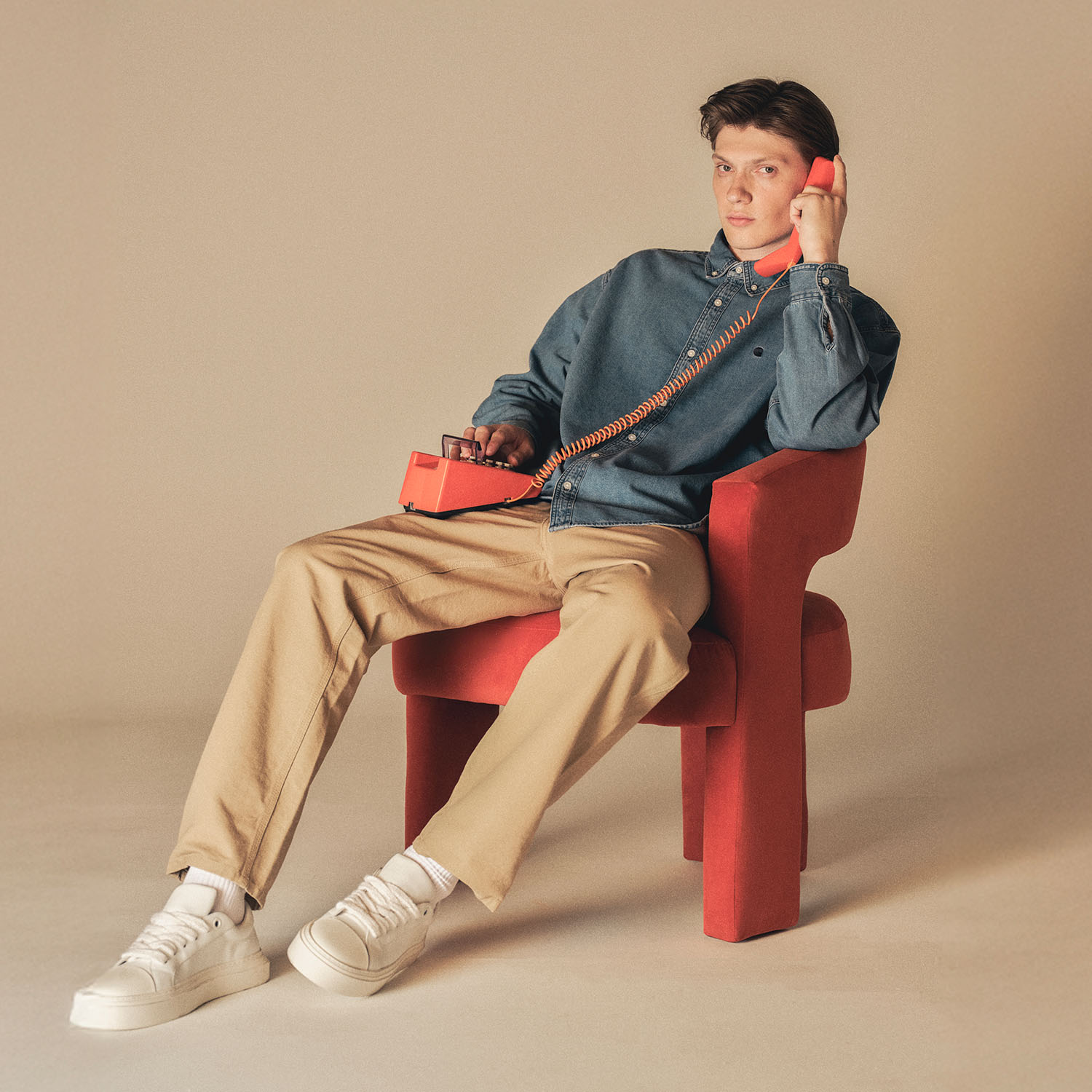 Person in casual attire sits on a red chair, holding a vintage red telephone to their ear.