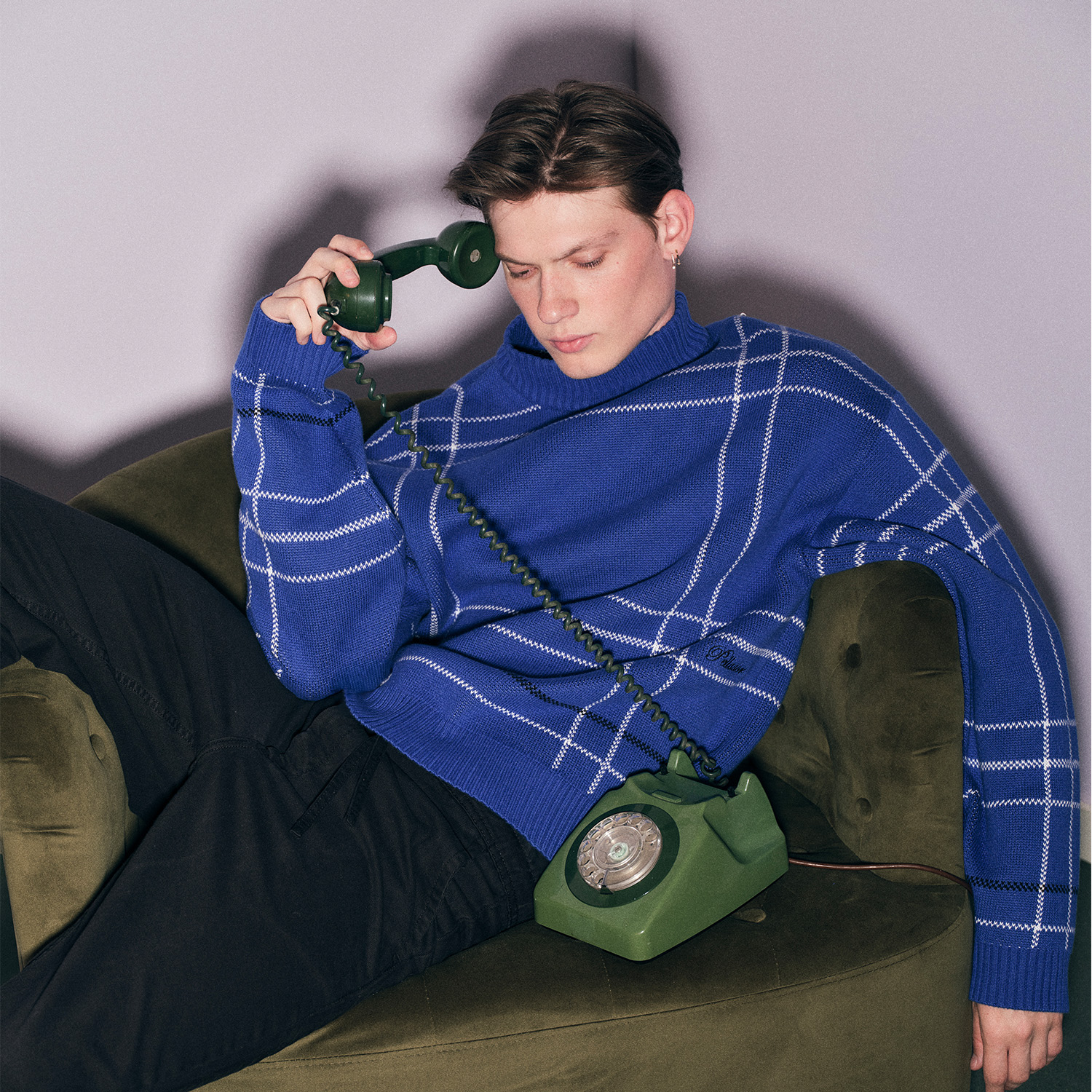 A person in a blue Pellador sweater lounges on a green chair, holding a vintage rotary phone to their ear. 