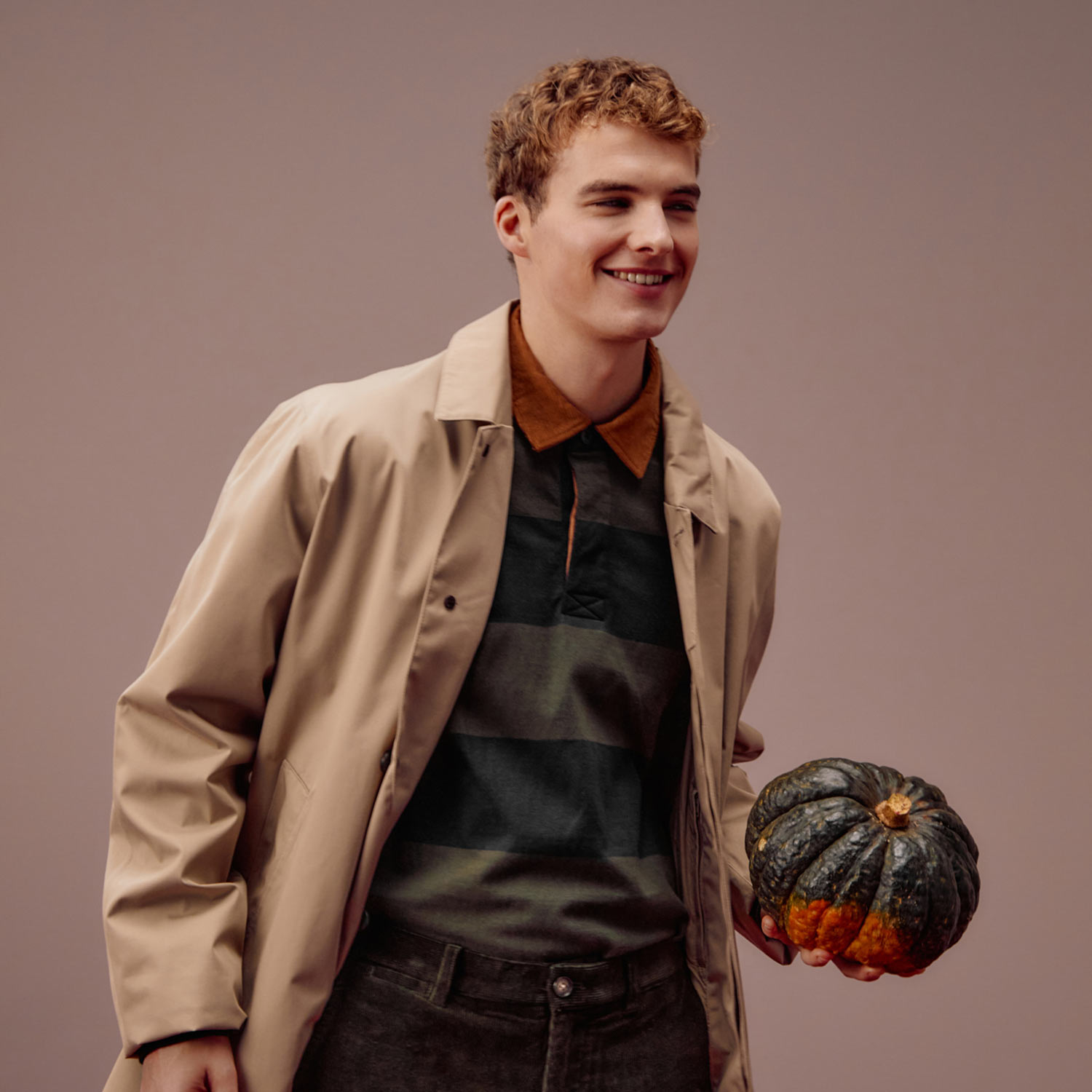 A smiling person in a beige coat holds a dark green pumpkin.