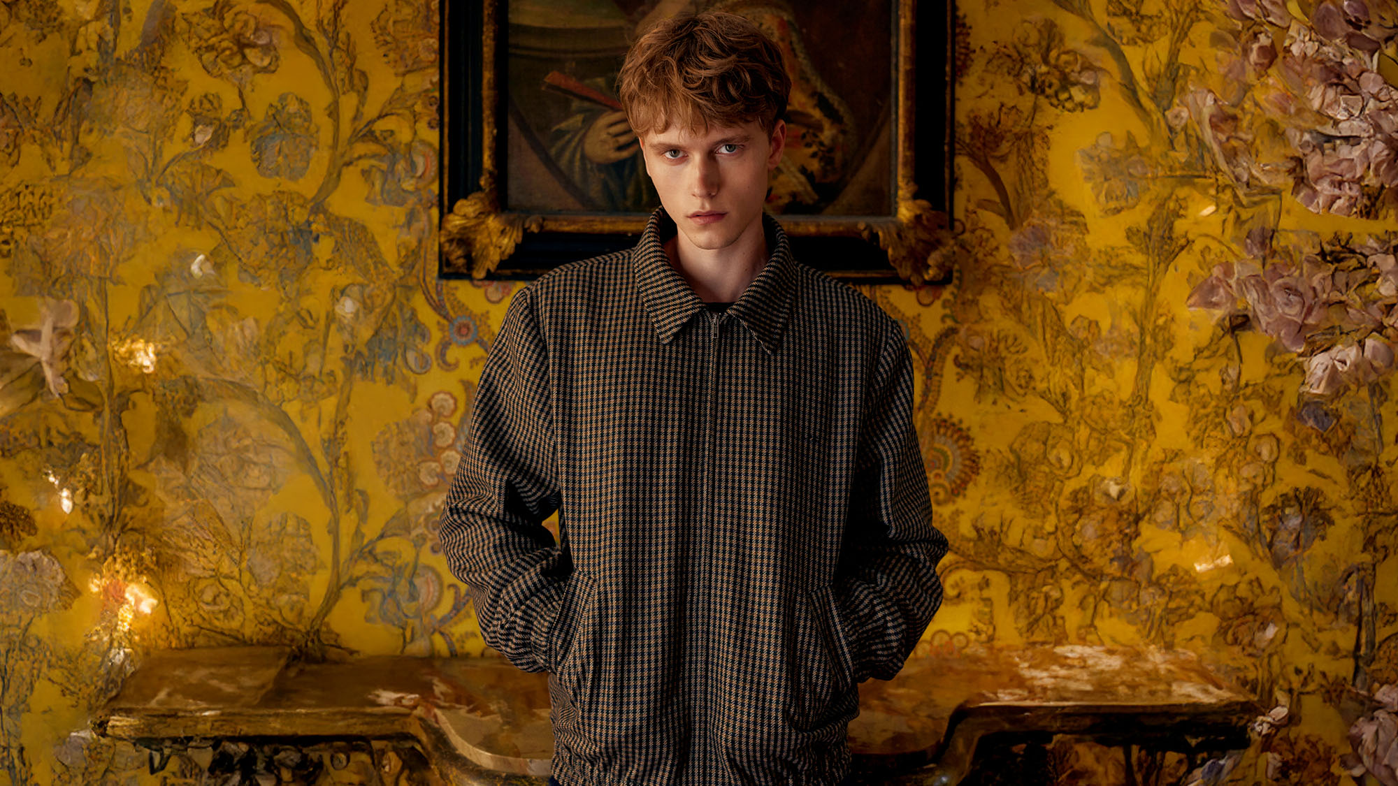 Young man in a patterned jacket stands in a vintage room with ornate wallpaper and a framed painting behind him.