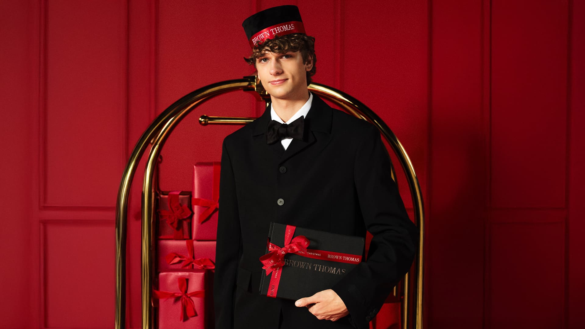 A person in formal attire holds a gift box in front of a red background with stacked presents.