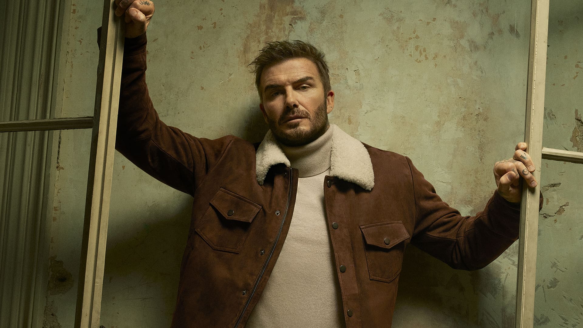 A man in a brown jacket with a shearling collar stands between open window frames, looking toward the camera.
