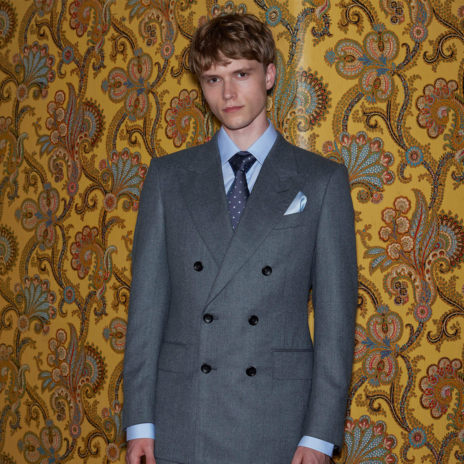 A person in a gray double-breasted suit stands against a floral-patterned yellow wallpaper.