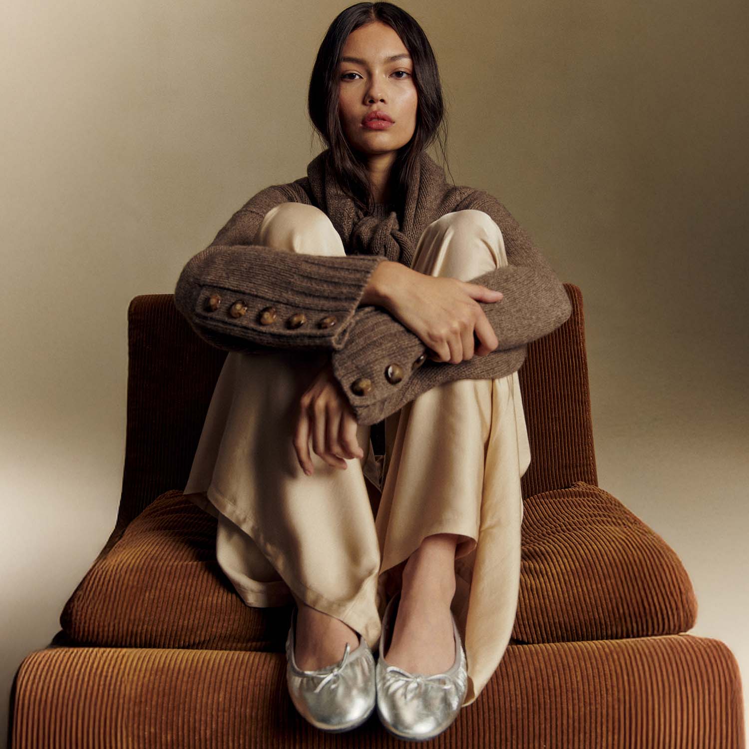 A person wearing a brown Sézane sweater, cream pants, and silver shoes sits on a chair. 